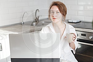 Happy beautiful woman with cup of coffee or tea using laptop in quarantine lockdown in the kitchen in the white shirt