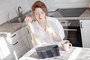 Happy beautiful woman with cup of coffee or tea using laptop in quarantine lockdown in the kitchen in the white shirt
