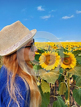 A happy, beautiful woman in a hat with sunglasses sits in a large field of sunflowers on a sunny summer day