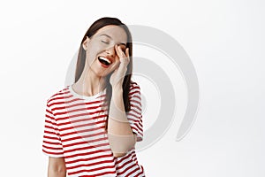 Happy beautiful woman laughing, touching face with closed eyes and tender face expression, smiling carefree, standing in