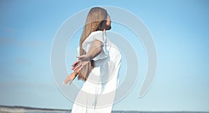 Happy, beautiful woman on the ocean beach standing in a white summer dress, open arms