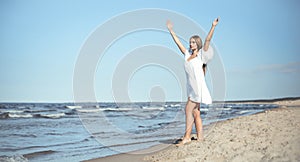 Happy, beautiful woman on the ocean beach standing in a white summer dress, raising hands