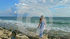 Happy beautiful woman standing on rocky sea beach, foamy waves crashing ashore