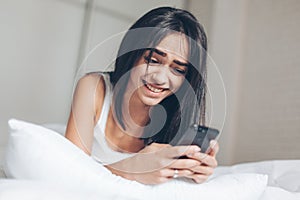 Happy Beautiful woman texting at smartphone while lying on her bed. Smiling Brunette Girl holding mobile phone