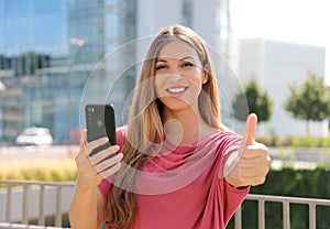 Happy beautiful woman thumbs up with smartphone in her hand and modern city on background. Positive businesswoman smiles at camera