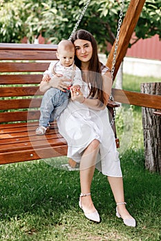 Happy beautiful woman, young mother playing with her adorable baby son and sitting on wooden swing, cute little boy