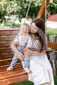 Happy beautiful woman, young mother playing with her adorable baby son and sitting on wooden swing, cute little boy