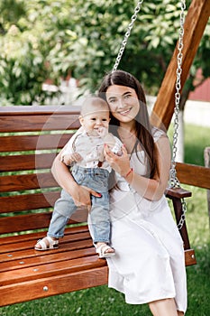 Happy beautiful woman, young mother playing with her adorable baby son and sitting on wooden swing, cute little boy
