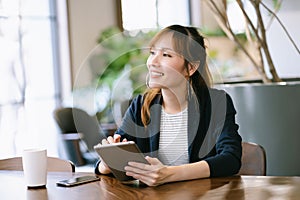 Happy beautiful Young Asian business woman holding digital tablet and looking away while sitting at table in co-working office.
