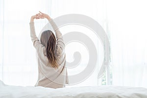 Happy beautiful young Asian woman waking up in morning, sitting on bed, stretching in cozy bedroom, looking through window.