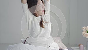 Happy beautiful young Asian woman waking up in morning, sitting on bed, stretching in cozy bedroom, looking through window.
