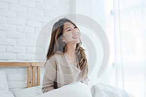 Happy beautiful young Asian woman waking up in morning, sitting on bed, stretching in cozy bedroom.