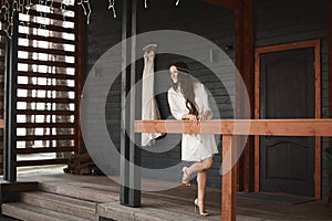 Happy and beautiful young brunette bride with wedding hairstyle and makeup, wearing nightwear, standing on a porch and