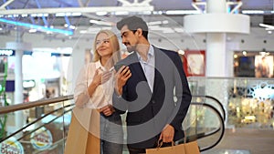 Happy beautiful young couple using mobile phone standing with paper bags with purchases in shopping mall.