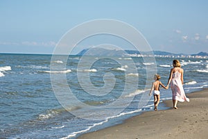 Happy beautiful young mom and child walking along the sea beach, waves and wind