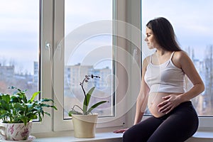 Happy beautiful young pregnant woman dreamily looking out home window