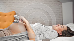 Happy beautiful young pregnant woman having fun playing with little cute baby shoes lying on bed