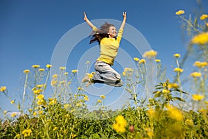 Happy and beautiful young woman in a bright yellow sweater and blue jeans jumping high in a sunny summer field