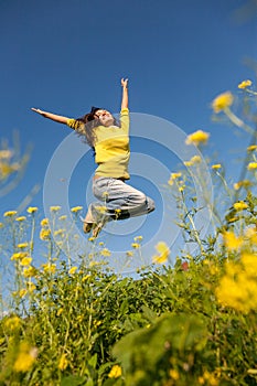 Happy and beautiful young woman in a bright yellow sweater and blue jeans jumping high in a sunny summer field