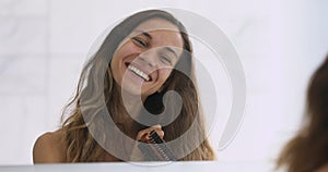 Happy beautiful young woman brushing long hair looking in mirror