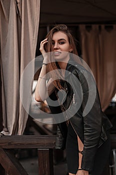 Happy beautiful young woman in a fashionable leather black jacket in a black elegant dress is resting in a vintage cafe. Joyful