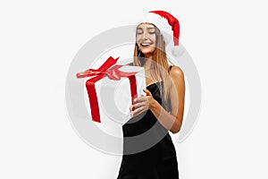 Happy beautiful young woman in a red Santa Claus hat and in a black elegant dress, with a gift on a white background, holiday