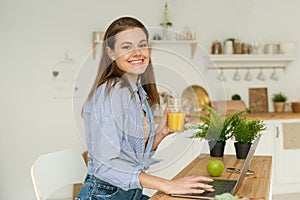 Happy beautiful young woman sitting at the table at home, working in a laptop and drinking delicious orange juice