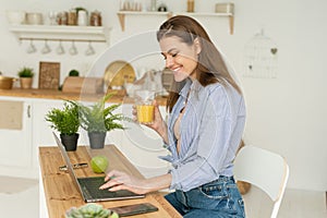 Happy beautiful young woman sitting at the table at home, working in a laptop and drinking delicious orange juice. Work