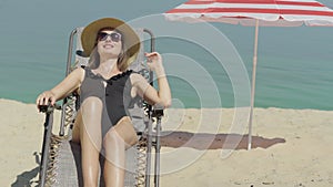 Happy beautiful young woman in swimsuit and sunglasses resting on sun lounger on sandy beach. Portrait of smiling