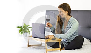 Happy Beautiful young woman teleworking at home in a bed with laptop and glass of red wine, lifestyle, copy space. Freelance and