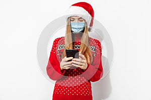 Happy young woman, wearing red santa claus hat and medical protective mask on face, with mobile phone, isolated on white