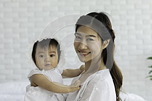 Happy Beauty mother Holding Cute Sweet Adorable Asian Baby wearing white dress sitting on Carpet smiling