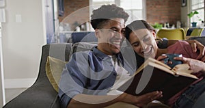 Happy biracial couple sitting on sofa in living room, reading book and drinking coffee