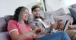 Happy biracial couple sitting on sofa and using tablet for online shopping at home, in slow motion