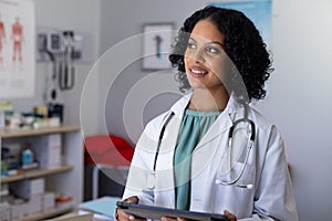 Happy biracial female doctor wearing sthethoscope, using tablet at doctor\'s office photo