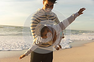 Happy biracial man carrying son pretending to fly with arms outstretched at beach during sunset