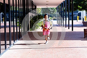 Happy biracial schoolgirl with school bag, holding books and running outside school, with copy space