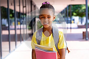 Happy biracial schoolgirl with school bag holding books, smiling outside school, with copy space
