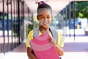 Happy biracial schoolgirl with school bag holding books, smiling outside school, with copy space