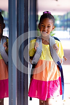 Happy biracial schoolgirl with school bag smiling in sun outside school, with copy space