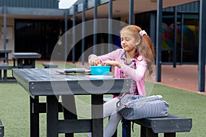 Happy biracial schoolgirl sitting at table with packed lunch in schoolyard