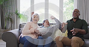 Happy biracial senior couple and african american friend watching tv with popcorn