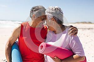 Happy biracial senior couple holding yoga mats touching foreheads of each other at beach