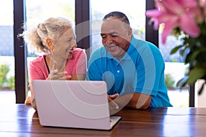 Happy biracial senior man and caucasian woman watching video over laptop while sitting at table