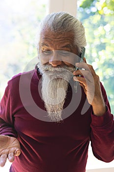 Happy biracial senior man with long beard, using smartphone in sunny living room, copy space