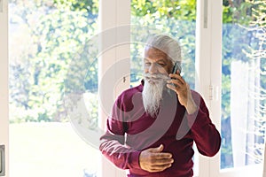 Happy biracial senior man with long beard, using smartphone in sunny living room, copy space