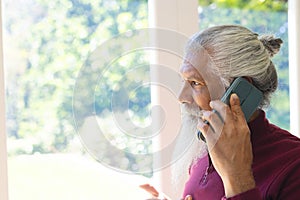 Happy biracial senior man with long beard, using smartphone in sunny living room, copy space