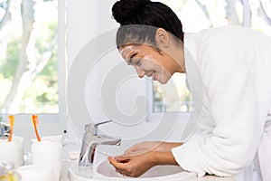 Happy biracial woman in bathrobe rinsing mask from her face in sunny bathroom