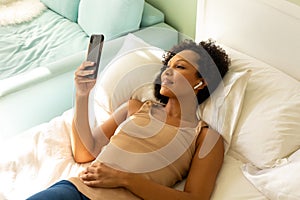 Happy biracial woman with earphones using smartphone and lying on bed in bedroom