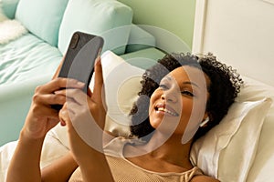 Happy biracial woman with earphones using smartphone and lying on bed in bedroom